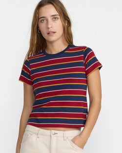 A woman wearing a short-sleeve crewneck t-shirt with horizontal stripes in navy blue, burgundy red, and mustard yellow, paired with cream-colored pants, against a plain white background. Brand RVCA,