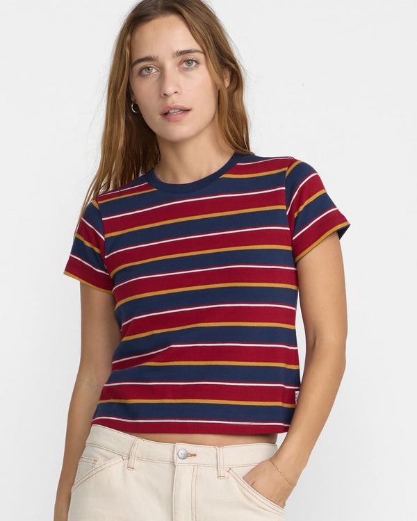 A woman wearing a short-sleeve crewneck t-shirt with horizontal stripes in navy blue, burgundy red, and mustard yellow, paired with cream-colored pants, against a plain white background. Brand RVCA,