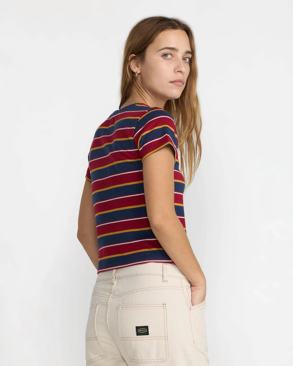 A woman wearing a short-sleeve striped crop top with red, navy, and yellow horizontal stripes, paired with off-white jeans, photographed against a plain white studio background. The brand is RVCA. Back to school style. Tee shirt style is called Unbound.